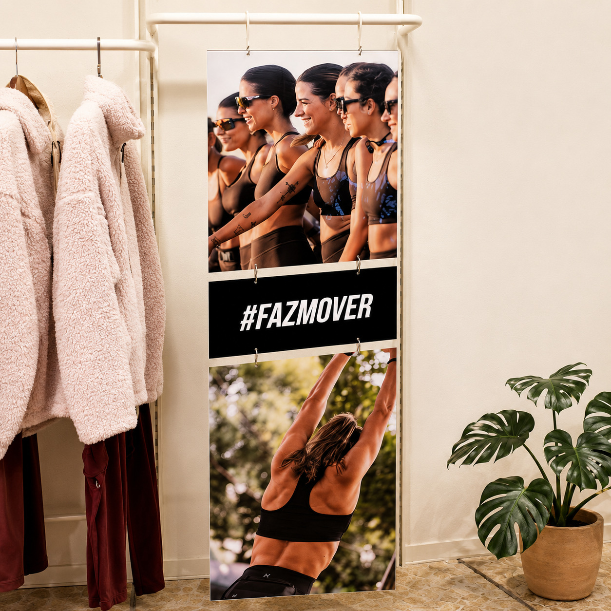 Banner de campanha publicitária com a hashtag '#FAZMOVER' e fotos de mulheres praticando exercícios, instalado em uma arara de loja ao lado de casacos peludos e um vaso com planta.