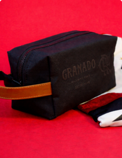 Nécessaire preta de tecido com alça lateral marrom em couro sintético e zíper superior, exibindo a personalização com as marcas 'Granado Pharmácias - Desde 1870' e o monograma do Flamengo.