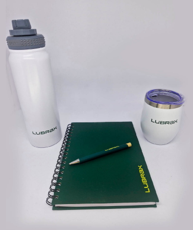 Kit de brindes corporativos da marca 'Lubrax' composto por uma garrafa de água branca com tampa cinza, um caderno com capa verde e espiral preta, uma caneta verde com detalhes amarelos e um copo térmico branco com tampa.