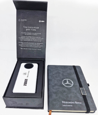 Kit de brindes corporativos de luxo em caixa preta com mensagem impressa na tampa interna, contendo um carregador portátil branco com as marcas 'Vibra' e 'Mercedes-Benz', ao lado de um caderno cinza texturizado com elástico preto e o logotipo da Mercedes-Benz.