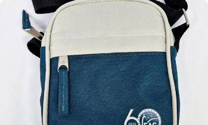 Bolsa transversal do tipo shoulder bag em tons de azul e bege, com zíper frontal e o logotipo comemorativo 