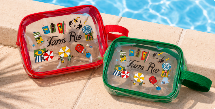 Kit de nécessaires transparentes da Farm Rio, uma vermelha e uma verde, com alças laterais e estampa de praia tropical, sobre a borda de uma piscina com água azul ao fundo.