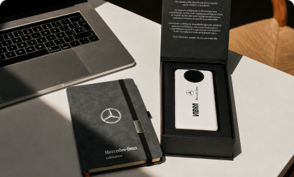 Kit corporativo personalizado disposto sobre uma mesa de escritório ao lado de um laptop, contendo um caderno com capa cinza da Mercedes-Benz Lubrificantes e uma caixa preta de presente com um carregador portátil co-branded das marcas Mercedes-Benz e Vibra.