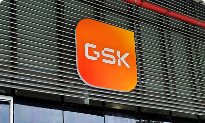 Letreiro corporativo da GSK com fundo em gradiente laranja e vermelho e logotipo em branco, instalado na fachada com brises metálicos de um edifício.