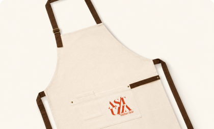 Avental de cozinha em tecido bege claro com alças e detalhes marrons, apresentando um bolso frontal com a logomarca da marca 