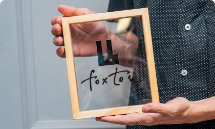 Pessoa segurando uma moldura de madeira com vidro transparente onde está impresso o logotipo e o nome da marca 'foxton' em preto.